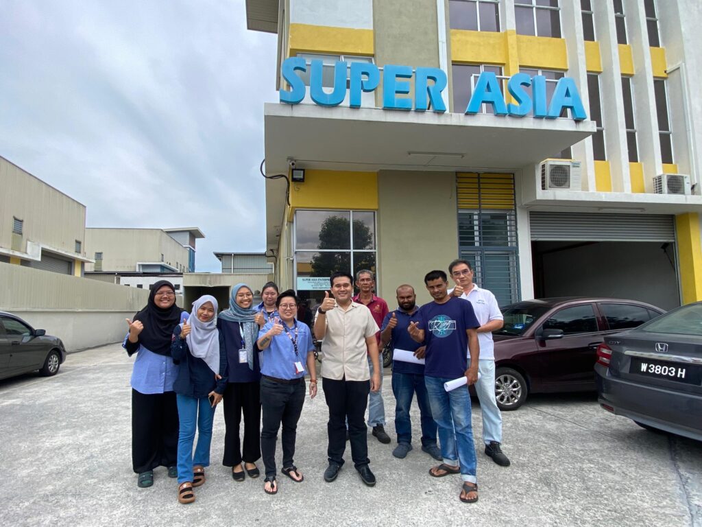 A group of diverse individuals standing in front of a building with the sign 'SUPER ASIA'. They are all smiling and giving thumbs up, showcasing a positive atmosphere.